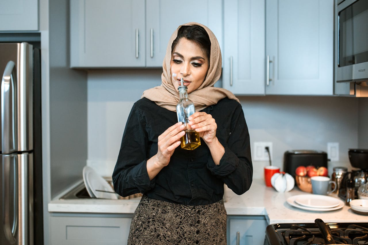 Positive stylish ethnic female in headscarf smelling aromatic olive oil in bottle while cooking dinner in contemporary light kitchen